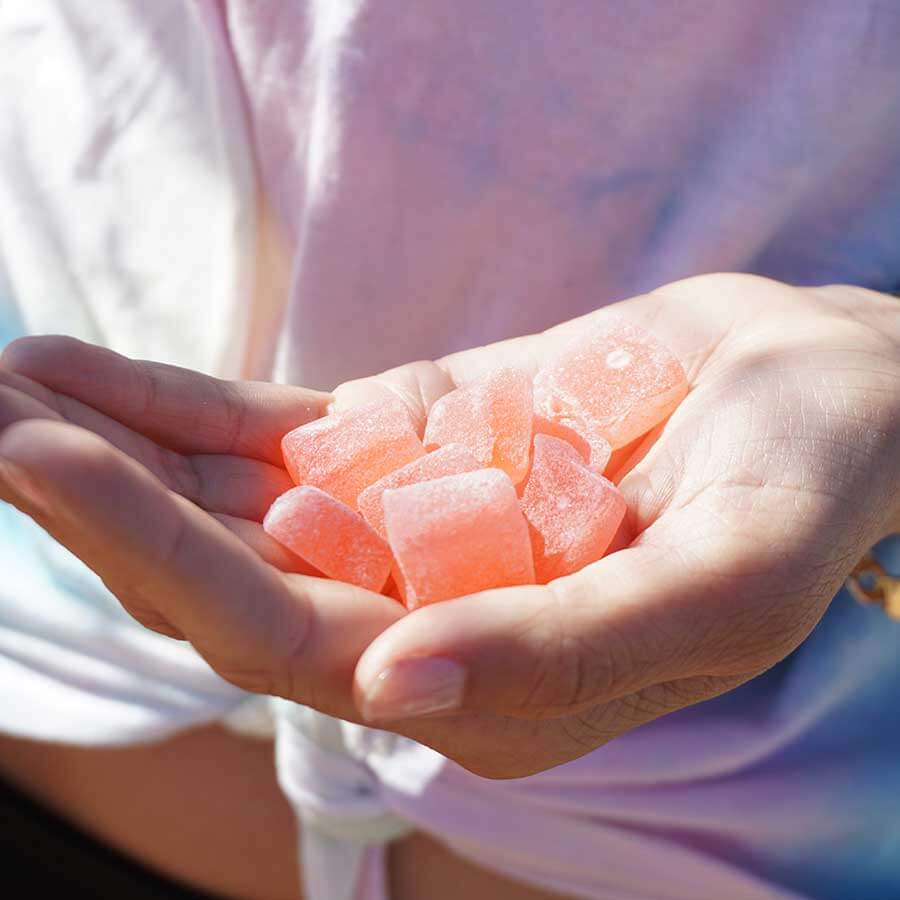 Close-up image of a person's hand holding several pink, translucent gummy candies in an outdoor setting.